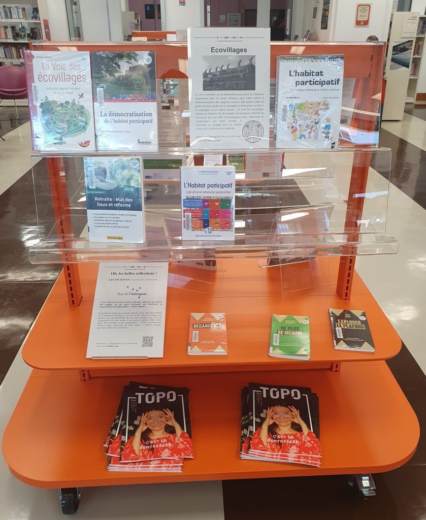 Une table de valorisation orange sur laquelle, en plus des ouvrages, se trouvent deux tas de Topo, et une affichette renvoyant vers L'Influx.