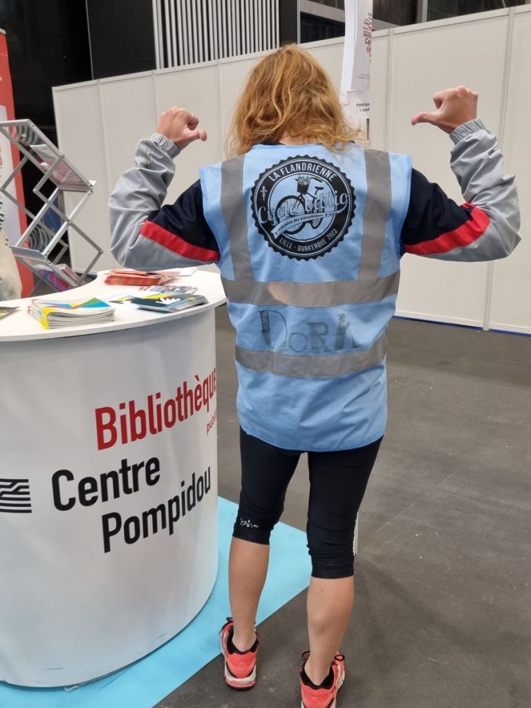Photo de Dora Blatigny de dos qui montre son gilet bleu de Cyclobiblio 2023 devant le stand Bpi du congrès ABF