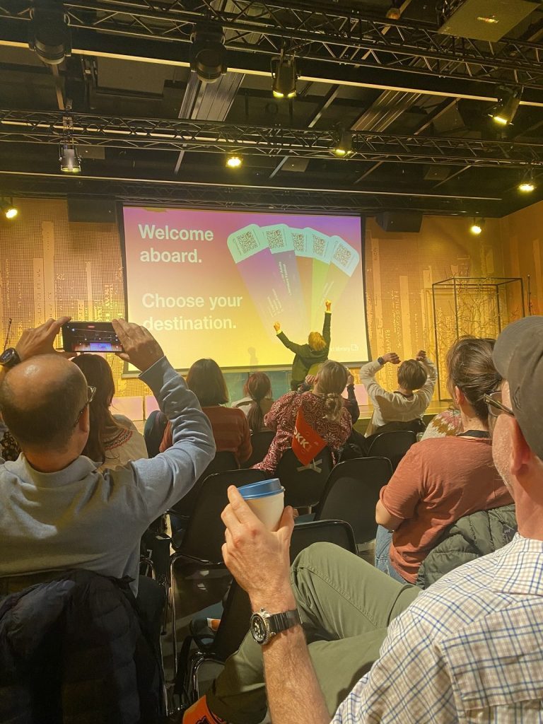 Une salle de conférence avec sur scène une femme de dos (la directrice de la bibliothèque) levant les bras et tout le monde dans la salle l'imitant.