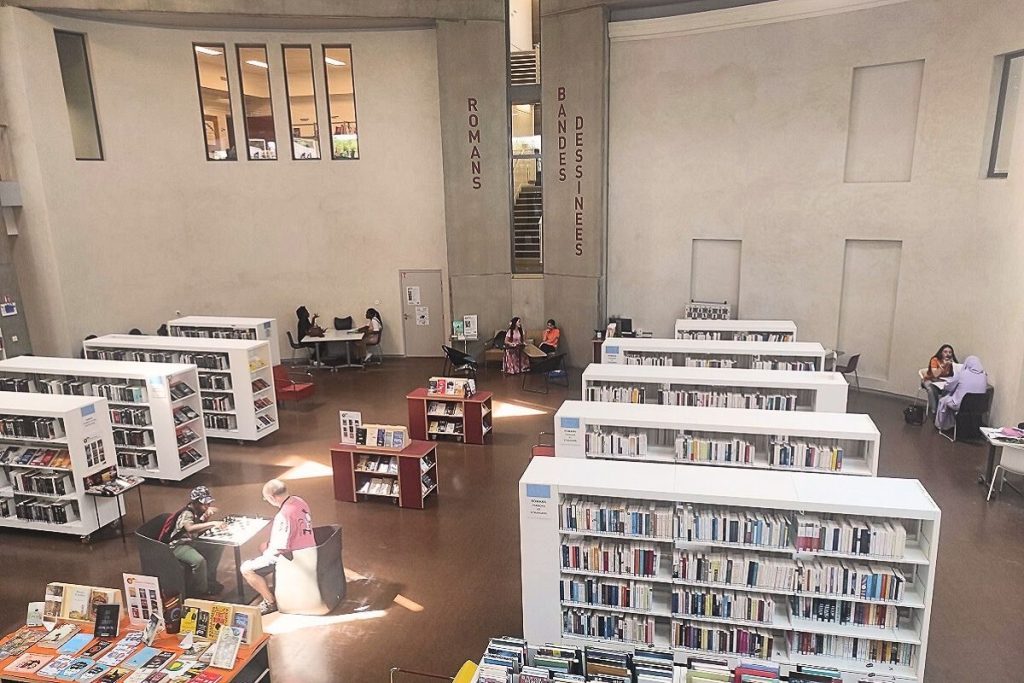 Espace interne de la médiathèque de Stains, grand espace ouvert avec des rayonnages et petites tables occupées par des personnes qui lisent, jouent aux échecs, etc.