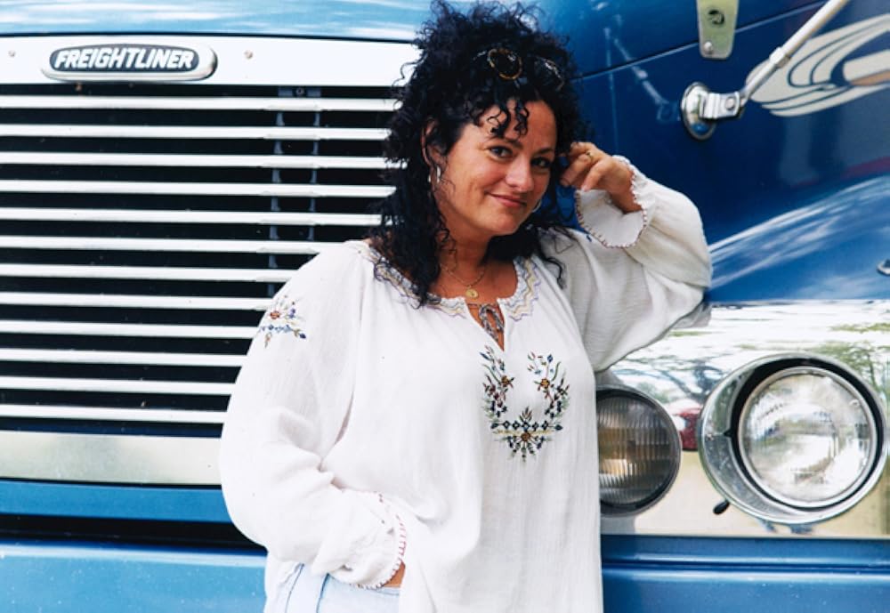 Une femme brune aux cheveux bouclés, dans une tunique blanche, pose en souriant devant un camion bleu.