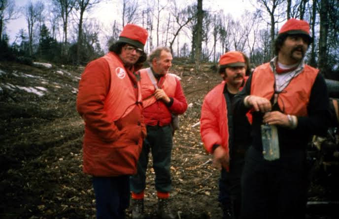 Quatre homme habillés en orange, dans une forêt, sourient à l'objectif.