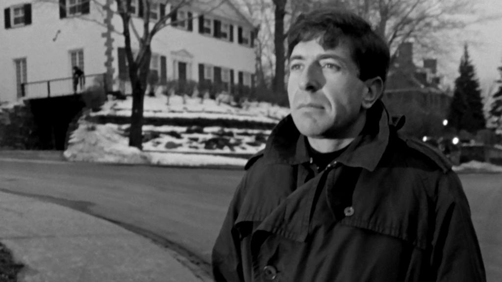 Léonard Cohen dans une rue enneigée, avec un manteau d'hiver, regardant au loin.