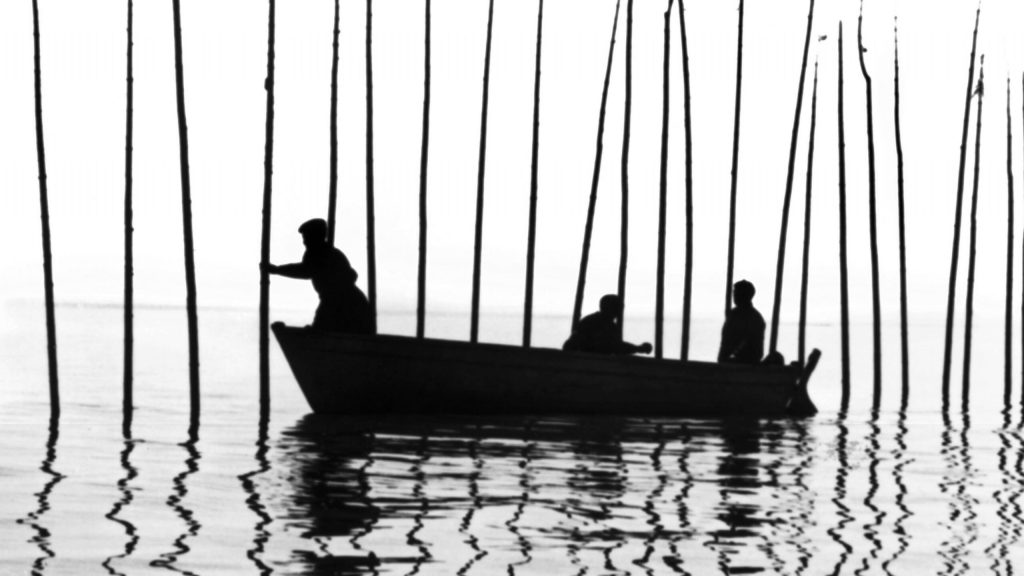 Trois personnes sur une barque, à contrejour, plantent dans l'eau de grands bâtons.
