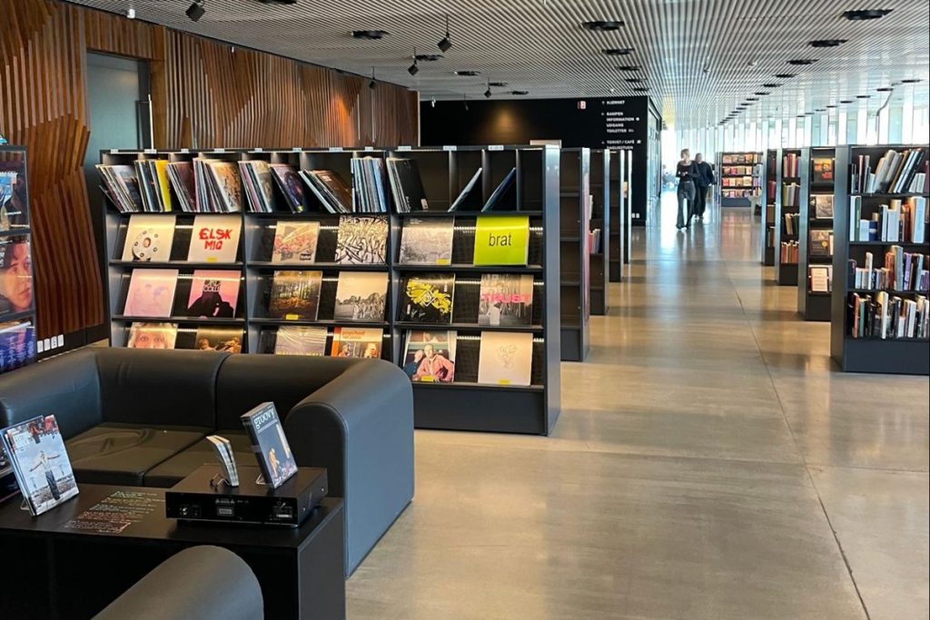 Des rayonnages noirs présentent des disques vinyles de face, dont certains avec des pochettes colorées reconnaissables. Au premier plan, un canapé en cuir gris et une table basse avec un lecteur CD. En arrière-plan, une longue allée entre d'autres rayonnages sous un plafond bas et des spots encastrés.