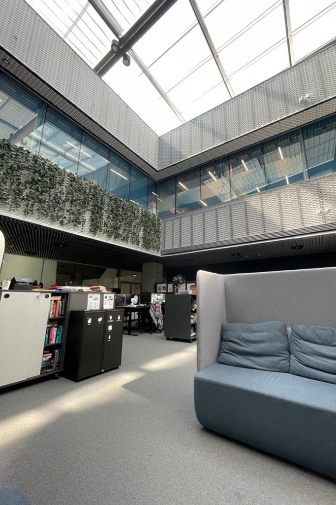 Un canapé gris à haut dossier est installé au premier plan. En arrière-plan, des rayonnages et des postes informatiques. Le bâtiment est surmonté d'une verrière zénithale, avec un mur végétal visible sur la mezzanine.