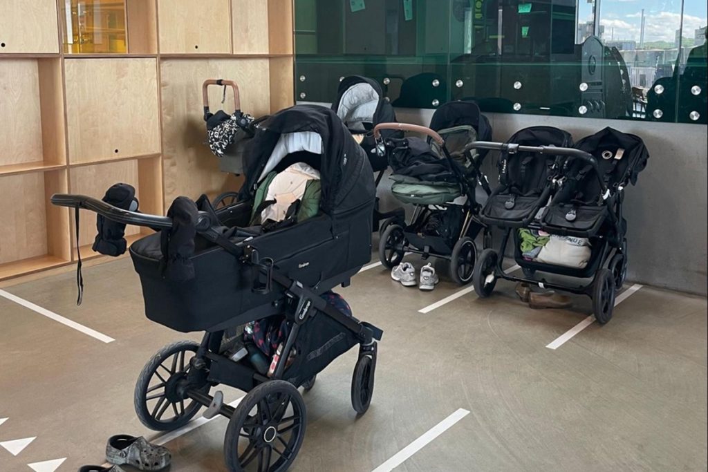 Espace de stationnement pour poussettes à l'entrée de la bibliothèque : quatre poussettes noires sont garées côte à côte dans des emplacements délimités par des lignes blanches au sol. Des chaussures sont posées à côté. Une paroi vitrée reflète l'extérieur.