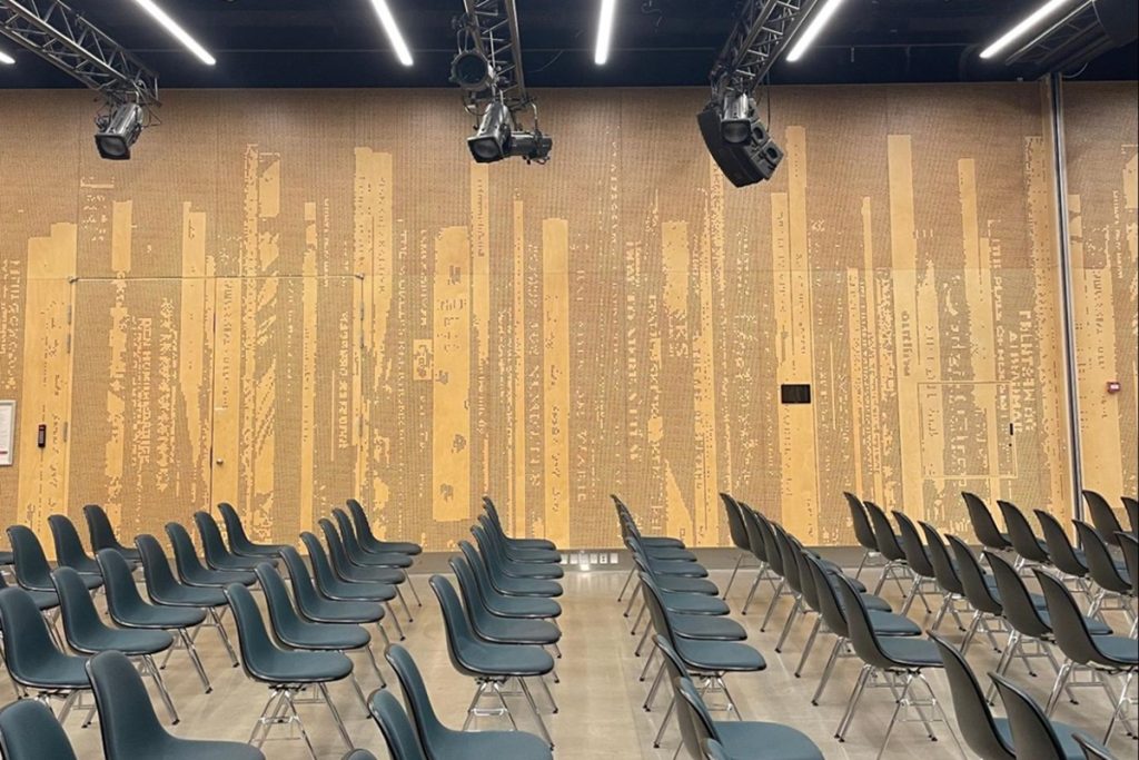 Grande salle de conférence vide, avec des rangées de chaises grises disposées en configuration théâtre. Le mur du fond est orné d'un grand panneau acoustique en relief beige, gravé de motifs représentant des dos de livres. Des projecteurs et des enceintes sont suspendus au plafond.