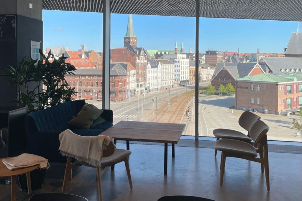 Espace avec des fauteuils et un canapé en velours bleu autour d'une table basse en bois. De larges baies vitrées offrent une vue panoramique sur le centre-ville d'Aarhus, avec ses toits en tuiles, ses bâtiments en briques et un clocher d'église sous un ciel bleu.
