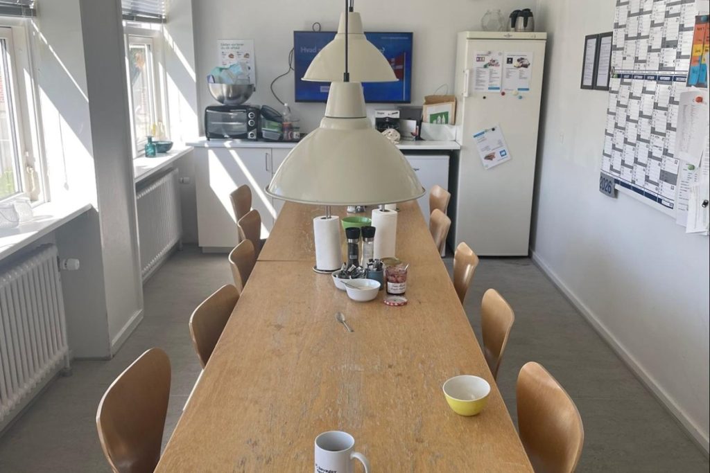 Une longue table en bois entourée de chaises en bois clair occupe le centre de la pièce. En fond, un coin cuisine avec réfrigérateur, micro-ondes et plan de travail. Sur le mur de droite, un calendrier et des affichages. Deux suspensions beiges éclairent la pièce.