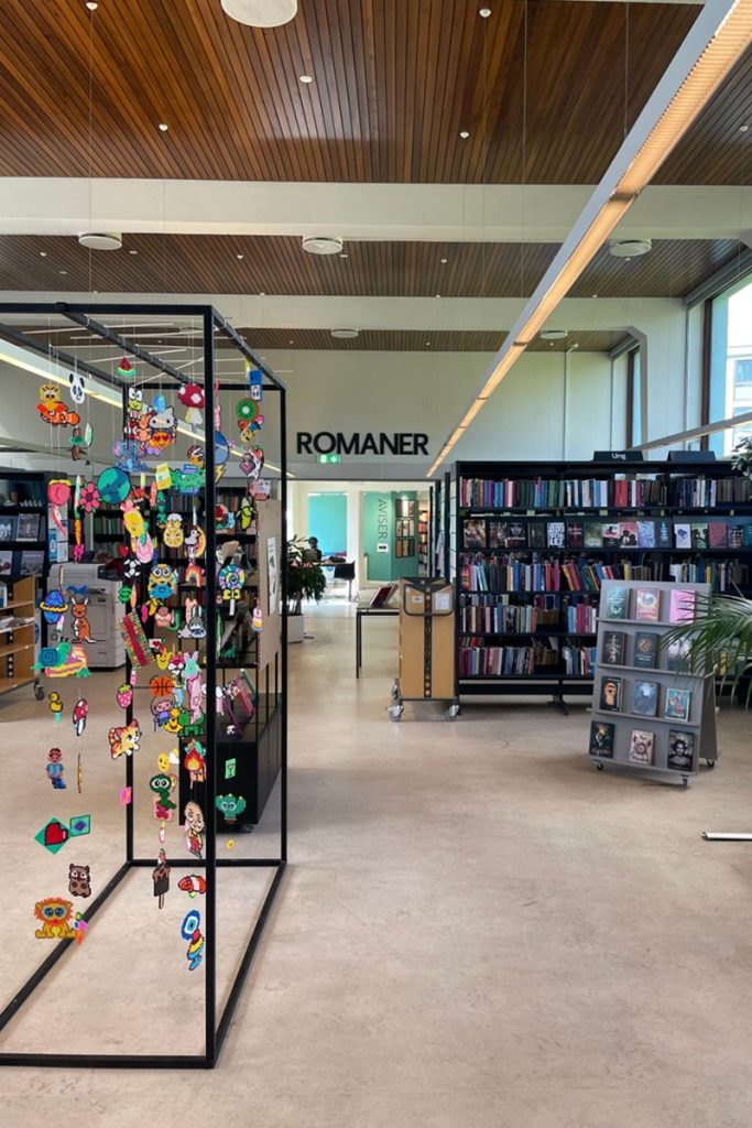 Au premier plan, une structure métallique noire présente en suspension de nombreuses figurines colorées en perles à repasser. En arrière-plan, des rayonnages de livres sous un panneau indiquant "Romaner" (romans en danois), avec un plafond en lattes de bois.