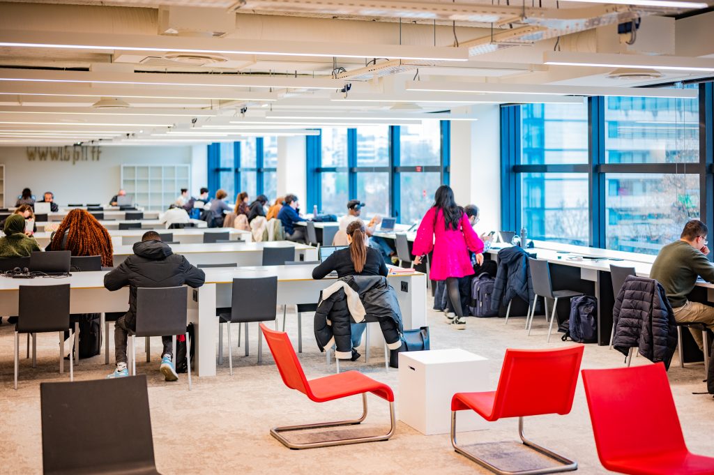 Vaste salle de travail animée par de nombreux usagers assis à de longues tables blanches, la plupart sur ordinateurs portables. Des chaises rouges en plastique sont disposées au premier plan. De larges baies vitrées à cadres bleus donnent sur l'extérieur.