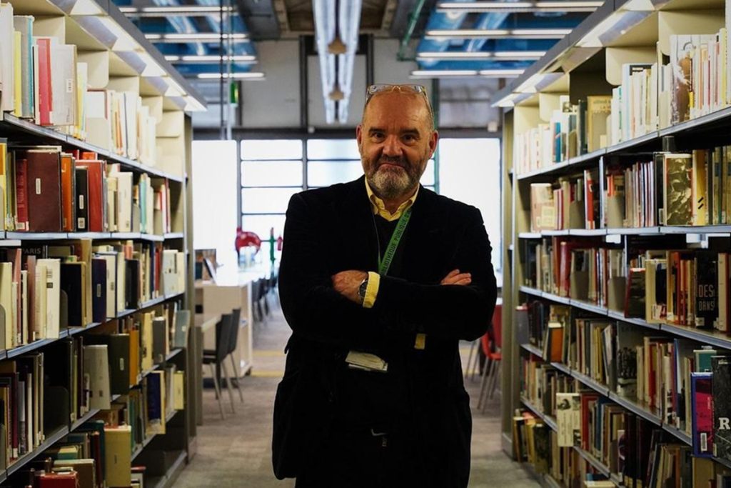 Portrait de Luis Cercós, debout les bras croisés entre deux rangées de rayonnages chargés de livres dans la bibliothèque du Centre Pompidou. Il porte un blazer noir et un badge vert autour du cou. La lumière provient d'une fenêtre en arrière-plan.