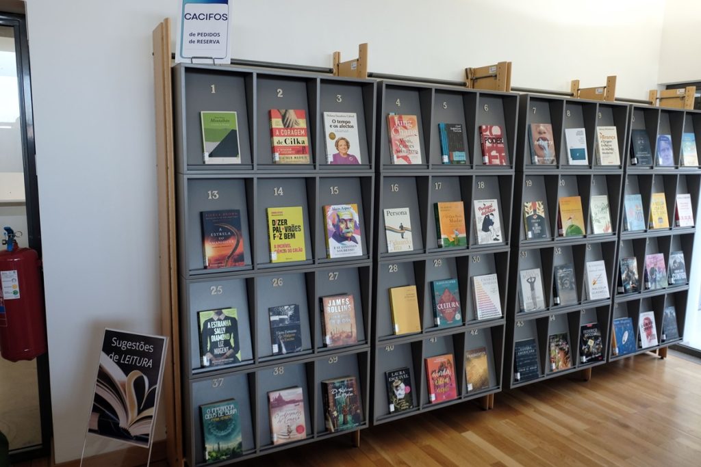 Mur de casiers numérotés en gris de la bibliothèque, chacun présentant un livre de face. Une pancarte en haut indique "Cacifos de pedidos de reserva" (casiers de réservation). Au premier plan à gauche, un présentoir "Sugestões de leitura" (suggestions de lecture).