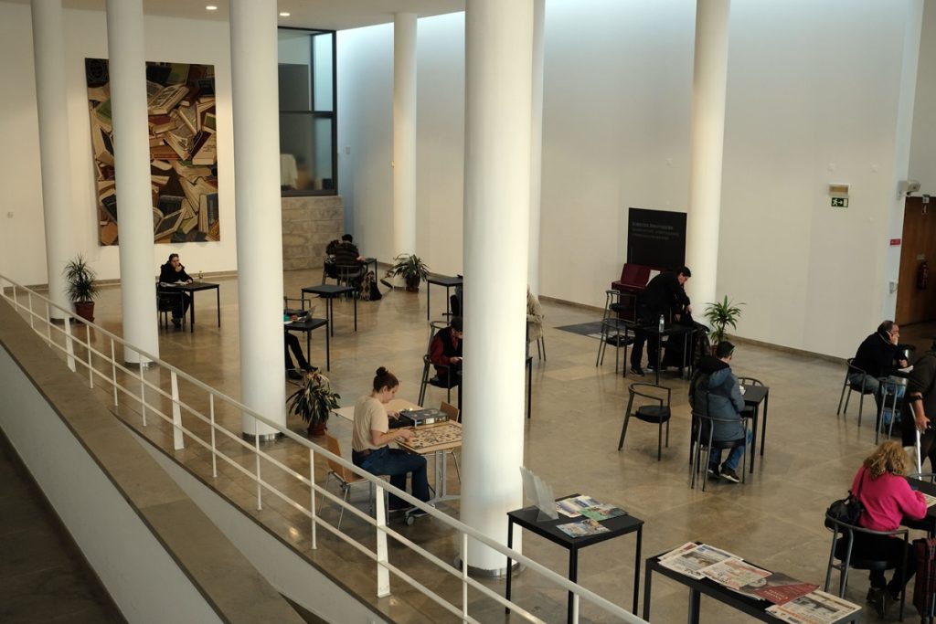 Hall spacieux de la bibliothèque vu depuis une mezzanine : de hautes colonnes blanches structurent l'espace lumineux. Plusieurs usagers sont assis seuls à de petites tables noires. Sur le mur de gauche, une grande peinture représente des livres empilés.