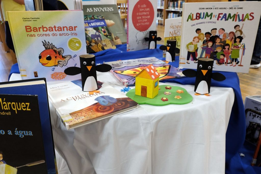 Table de présentation jeunesse : plusieurs livres pour enfants sont présentés debout sur une nappe blanc et bleu, dont "Barbatanar nas cores do arco-íris" et "Álbum de Famílias". Au centre, des figurines de pingouins en rouleaux de carton et une petite maison en carton coloré complètent la mise en scène.