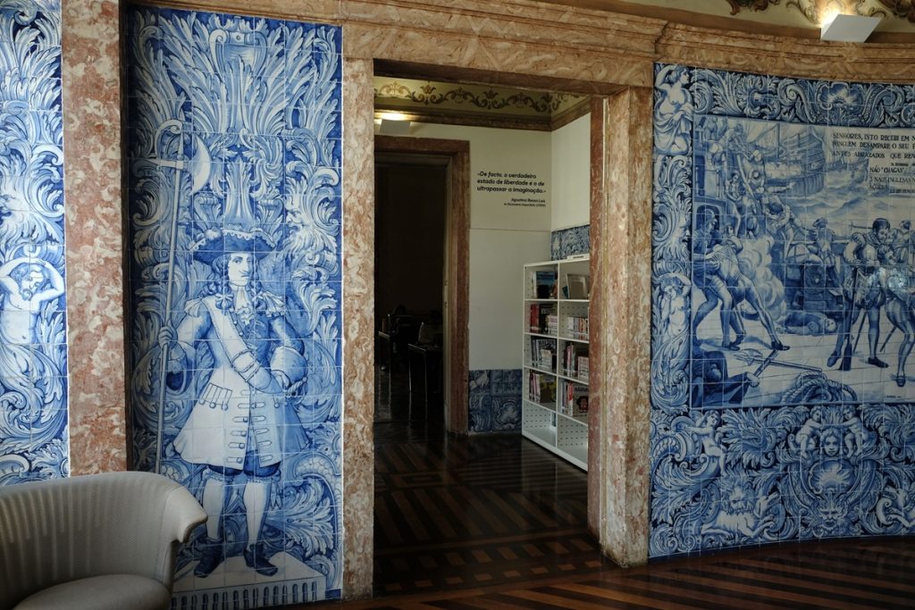 Intérieur de la bibliothèque : deux grands panneaux d'azulejos bleu et blanc encadrent une porte en marbre rose. Celui de gauche représente un personnage en habit d'époque, celui de droite une scène animée avec plusieurs personnages. En arrière-plan, une salle avec des rayonnages blancs et une citation murale.