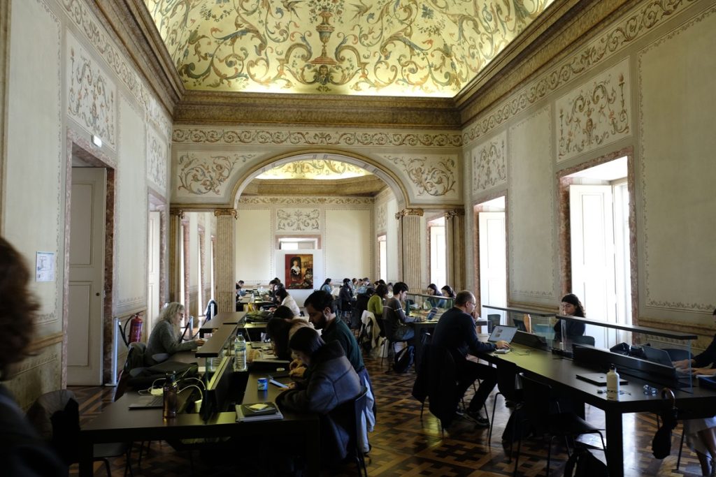 Salle de lecture de la bibliothèque : une pièce ornée d'un plafond à fresques florales en trompe-l'œil et de moulures dorées. De nombreux usagers travaillent sur des ordinateurs portables assis à longues tables sombres. Le parquet en marqueterie et les colonnes en marbre rose soulignent le caractère patrimonial du lieu.