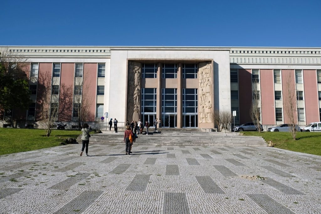Façade principale de la bibliothèque nationale : un imposant bâtiment de style moderniste en pierre beige et façades rose pâle, orné de deux hauts bas-reliefs sculptés encadrant l'entrée centrale. Un large parvis en pavés calcaires mène aux marches d'accès. Quelques visiteurs se trouvent devant l'entrée sous un ciel bleu dégagé.