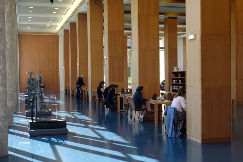 Grande salle de lecture : de hautes colonnes en bois bordent un vaste espace lumineux au sol bleu. Plusieurs usagers sont assis à des tables individuelles, travaillant sur ordinateurs portables ou lisant. À gauche, une ancienne machine d'imprimerie est exposée sur un socle.