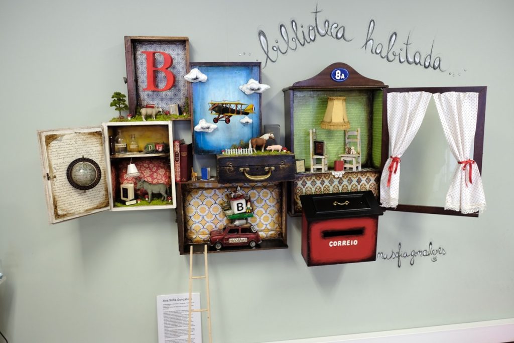 Œuvre artistique murale intitulée "biblioteca habitada" (bibliothèque habitée), signée Ana Sofia Gonçalves. L'installation est composée de plusieurs boîtes et objets assemblés sur un mur bleu pâle : une valise, une armoire miniature, une fenêtre avec des rideaux blancs, une boîte aux lettres rouge et divers éléments miniatures évoquant des scènes de vie et des univers imaginaires.