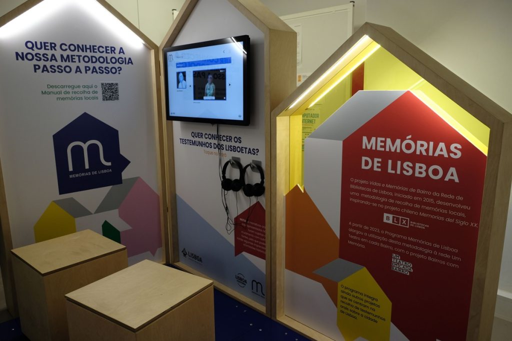 Le dispositif de médiation du projet "Memórias de Lisboa" se compose de trois panneaux en forme de maison en bois : celui de gauche présente la méthodologie du projet avec un QR code, celui du centre intègre un écran vidéo et un casque audio pour écouter des témoignages des habitants, celui de droite, en rouge, présente le programme et son lien avec le réseau des bibliothèques de Lisbonne.