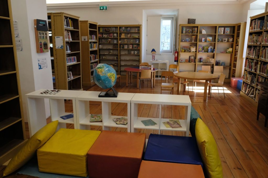 Espace jeunesse de la bibliothèque : au premier plan, des poufs colorés (jaune, orange, bleu) et des étagères basses avec un globe terrestre. En arrière-plan, des rayonnages en bois garnis de livres pour enfants, des tables et chaises adaptées, sur un parquet en bois clair.