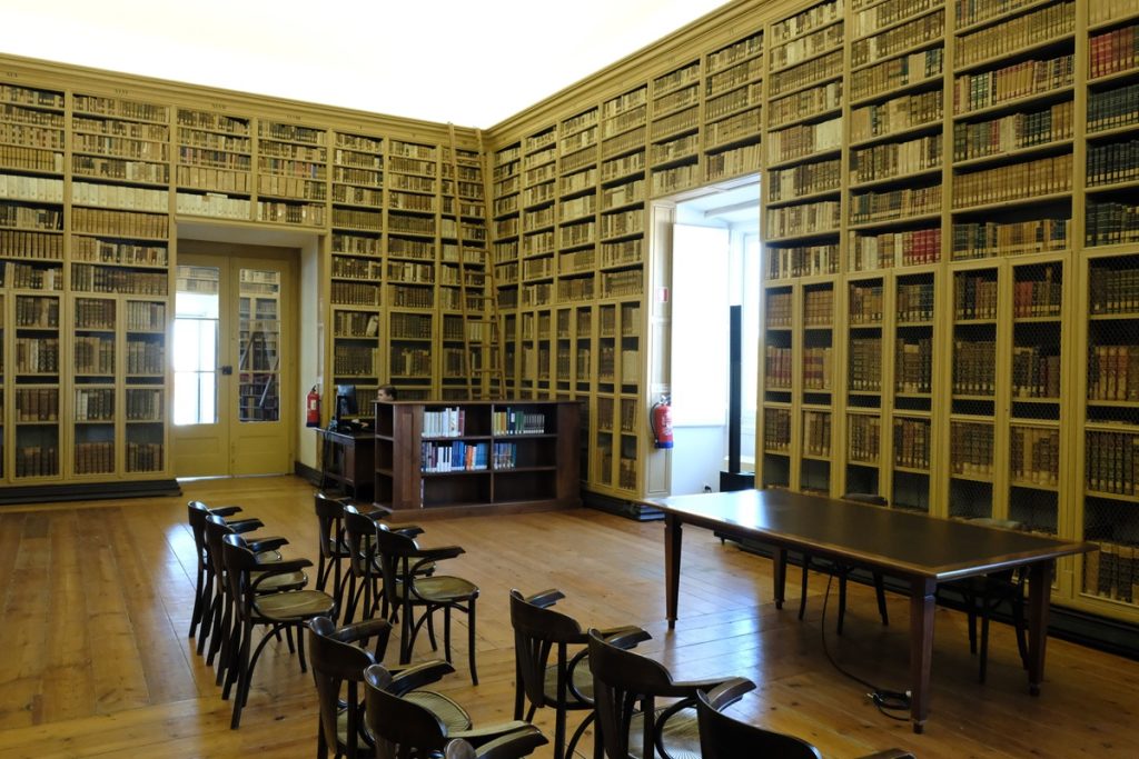 De hauts rayonnages en bois clair chargés d'ouvrages anciens couvrent entièrement les murs sur plusieurs niveaux. Au centre de la pièce, des chaises en bois sombre sont disposées autour de tables. Le parquet en bois clair et les fenêtres à volets blancs confèrent à l'espace une atmosphère classique et lumineuse.