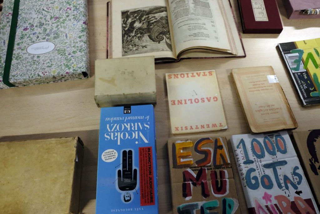 Vue en plongée sur une table couverte de livres et documents variés : un ouvrage ancien ouvert avec une gravure, un carnet à motifs floraux, plusieurs livres aux couvertures colorées, parmi lesquels "Gasoline Stations Twentysix" et "Le manuel vaudou" de Nicolas Sarkozy.