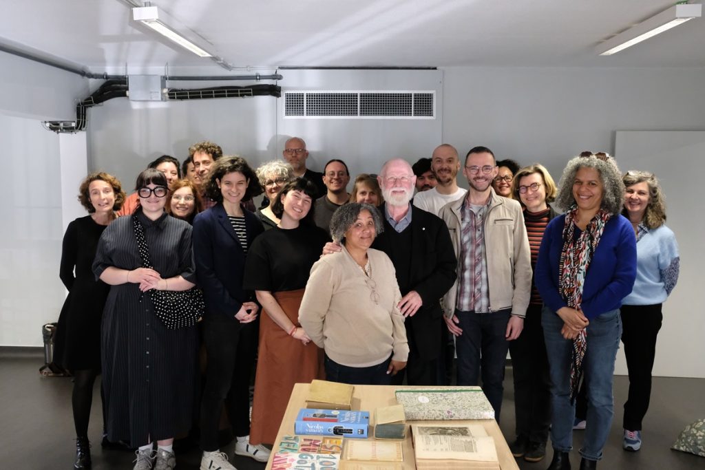Photo de groupe d'une vingtaine de personnes souriantes réunies autour de l'écrivain Alberto Manguel. Au premier plan, une table présente plusieurs livres issus de sa bibliothèque.