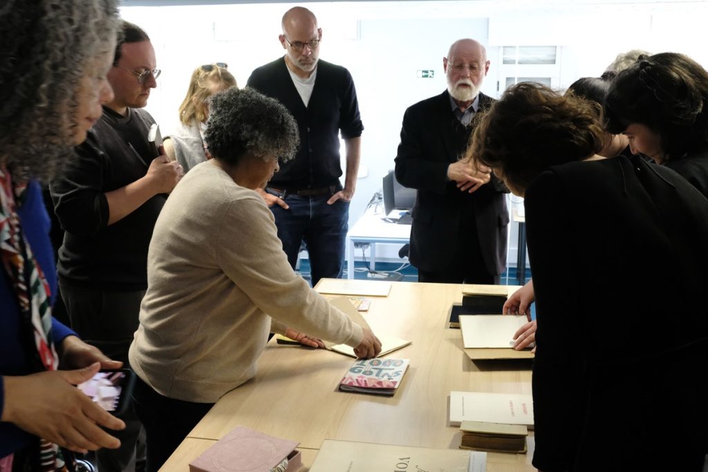 Un groupe d'une dizaine de personnes est réuni autour d'une table sur laquelle sont disposés plusieurs livres ouverts et des documents. Une femme en pull beige manipule l'un des ouvrages, tandis que les autres participants l'observent attentivement. L'écrivain Alberto Manguel se tient debout face au groupe.