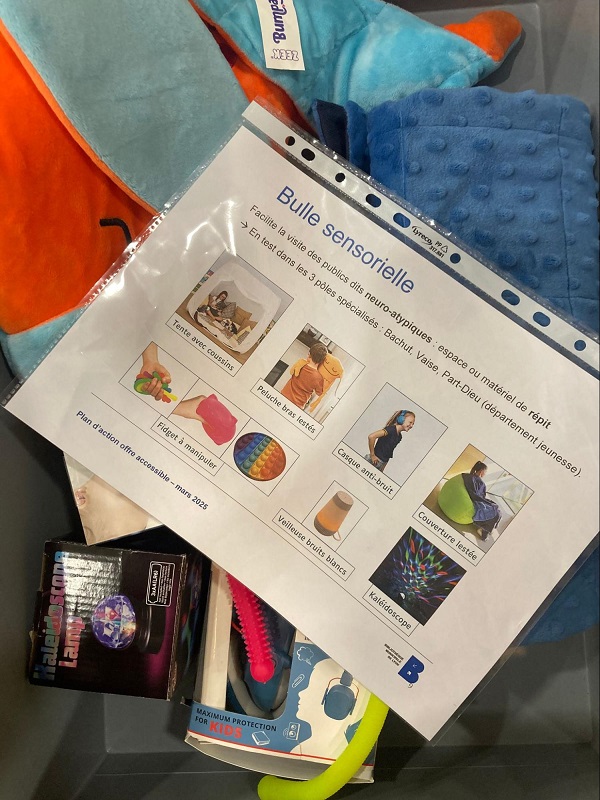 Vue en plongée sur un kit "Bulle sensorielle" de la bibliothèque. Une fiche plastifiée présente les différents objets du kit destinés aux publics neuroatypiques : tente avec coussins, peluche bras lestés, fidget à manipuler, casque anti-bruit, veilleuse bruit blanc, couverture lestée et kaléidoscope. Autour de la fiche, on aperçoit des coussins colorés et une boîte de kaléidoscope lumineux.