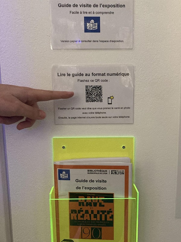 Dispositif d'accessibilité apposé sur un mur de la bibliothèque pour l'exposition "Entre rave et réalité". Deux panneaux indiquent la disponibilité d'un guide de visite "Facile à lire et à comprendre" (FALC) en version papier et numérique via QR code. Un présentoir jaune fluorescent contient les exemplaires papier du guide. Une main pointe le QR code.