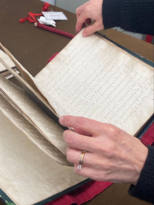 Deux mains feuilletant délicatement les pages d'un ancien ouvrage en braille, posé sur une table. Les pages en papier épais laissent apparaître les points en relief caractéristiques de l'écriture braille. En arrière-plan, d'autres documents et une étiquette rouge sont visibles.