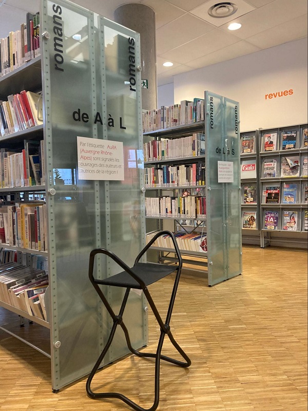 Espace romans de la bibliothèque : deux panneaux de signalétique en verre translucide indiquent "romans de A à L" et "romans de L à S". Un siège-canne pliable noir est disposé librement au pied du premier panneau, à disposition des usagers. En arrière-plan, des rayonnages de romans et un espace "revues".