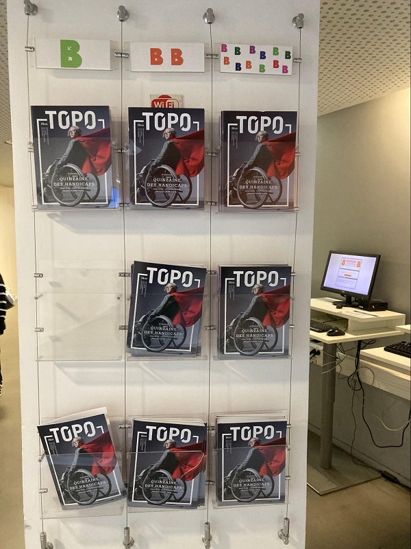 Présentoir mural transparent présentant neuf exemplaires du magazine "Topo", numéro spécial "Quinzaine des handicaps". La couverture représente une personne en fauteuil roulant en mouvement, drapée dans un tissu rouge. En haut du présentoir, des étiquettes de couleur indiquent les différents niveaux de lecture (B, BB, et plusieurs B de couleurs variées).