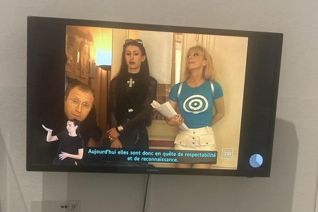 Écran de télévision accroché au mur diffusant une vidéo de l'INA sous-titrée et interprétée en langue des signes française (LSF). Une interprète en LSF est incrustée en bas à gauche de l'image. Un sous-titre indique : "Aujourd'hui elles sont donc en quête de respectabilité et de reconnaissance." En bas de l'écran, des pictogrammes indiquent la disponibilité de l'interprétation en LSF et du sous-titrage.