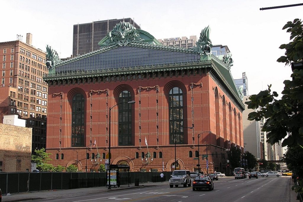 Façade de la Harold Washington Library à Chicago, vue depuis la rue. Le bâtiment en brique rouge foncé de style postmoderne est orné de grandes fenêtres cintrées et couronné d'un toit en verre bleu-vert avec des sculptures végétales monumentales en bronze patiné aux angles. La rue est animée par la circulation.