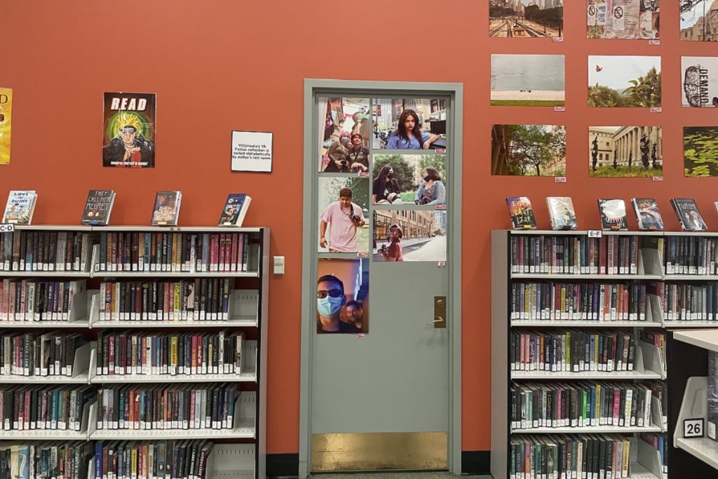 Espace YOUmedia dédié aux adolescents : deux rayonnages de romans face à une porte sur laquelle sont affichées des photos de jeunes usagers. Sur le mur orange, des photographies réalisées par des adolescents sont épinglées, avec une affiche "READ" à gauche. La cote indique un classement alphabétique par auteur.