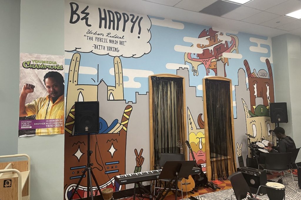 Espace musique de YOUmedia : une grande fresque murale colorée représente un paysage urbain stylisé avec la mention "Be Happy! The public needs art – Keith Haring". Des instruments de musique (clavier, guitare, djembé) et des enceintes sont installés devant la fresque. À gauche, une affiche "YOUmedia Champions" rend hommage à un ancien animateur.
