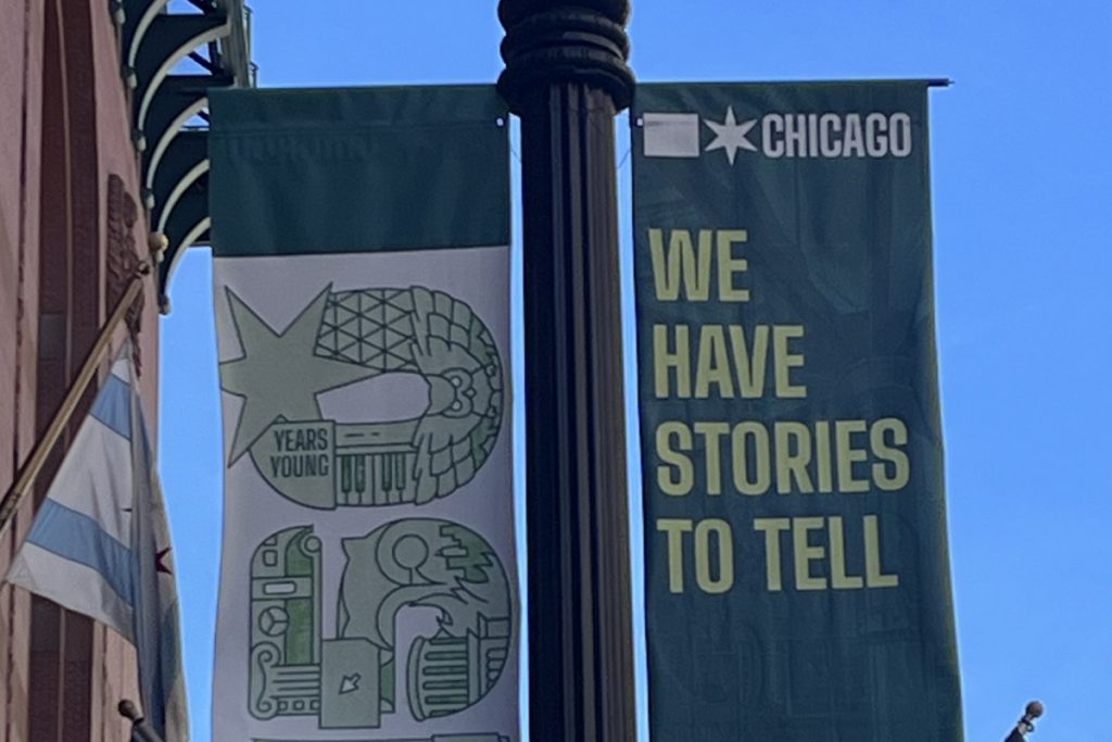 Deux bannières accrochées à un lampadaire devant la Harold Washington Library à Chicago : l'une, blanche, illustre le logo anniversaire des bibliothèques publiques de Chicago avec la mention "Years Young" ; l'autre, verte foncé, porte le slogan "Chicago – We have stories to tell" (Nous avons des histoires à raconter).