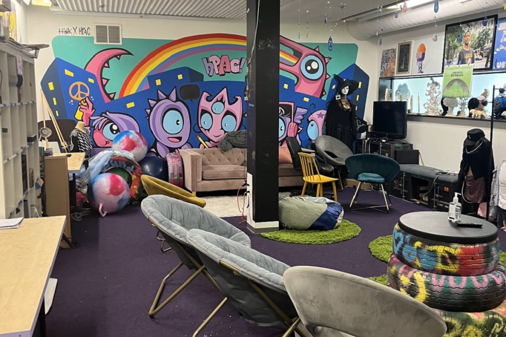 Espace dédié aux adolescents du Hyde Park Art Center : une grande salle au sol violet avec un mobilier varié et non conventionnel — fauteuils coquille, canapé, pneus colorés recyclés en tables, poufs. Le mur du fond est orné d'une grande fresque colorée aux personnages fantaisistes avec la mention "HPAC" et un arc-en-ciel. Des mannequins et des œuvres des jeunes sont exposés sur une étagère.