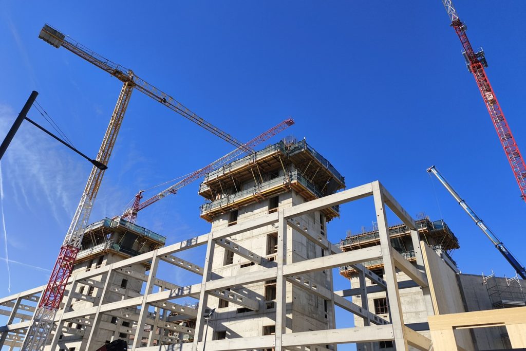 Vue en contre-plongée du chantier de la BEIC à Milan. Une structure en béton brut de plusieurs étages est en cours d'élévation, encadrée par trois grues de chantier sous un ciel bleu dégagé.