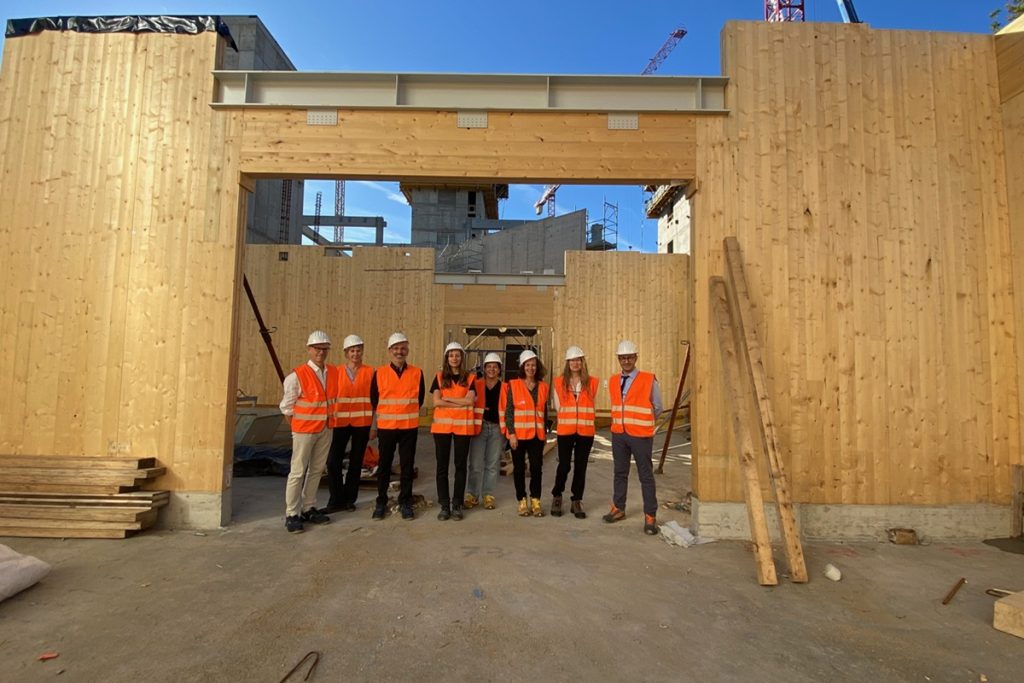 Groupe de huit personnes en gilets orange et casques blancs de sécurité posant sur le chantier de la BEIC, devant une grande structure en bois lamellé-croisé formant un portique. En arrière-plan, la structure en béton du bâtiment en construction et des grues sont visibles sous un ciel bleu.