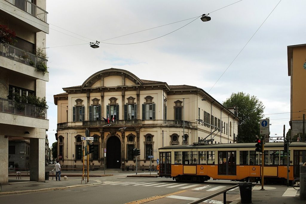 Façade du Palais Sormani, vue depuis la rue. Le bâtiment de style néoclassique arbore un fronton arrondi et des fenêtres ornementées. Au premier plan, un tramway orange caractéristique de Milan traverse l'intersection. Des drapeaux italien et européen sont visibles sur la façade.
