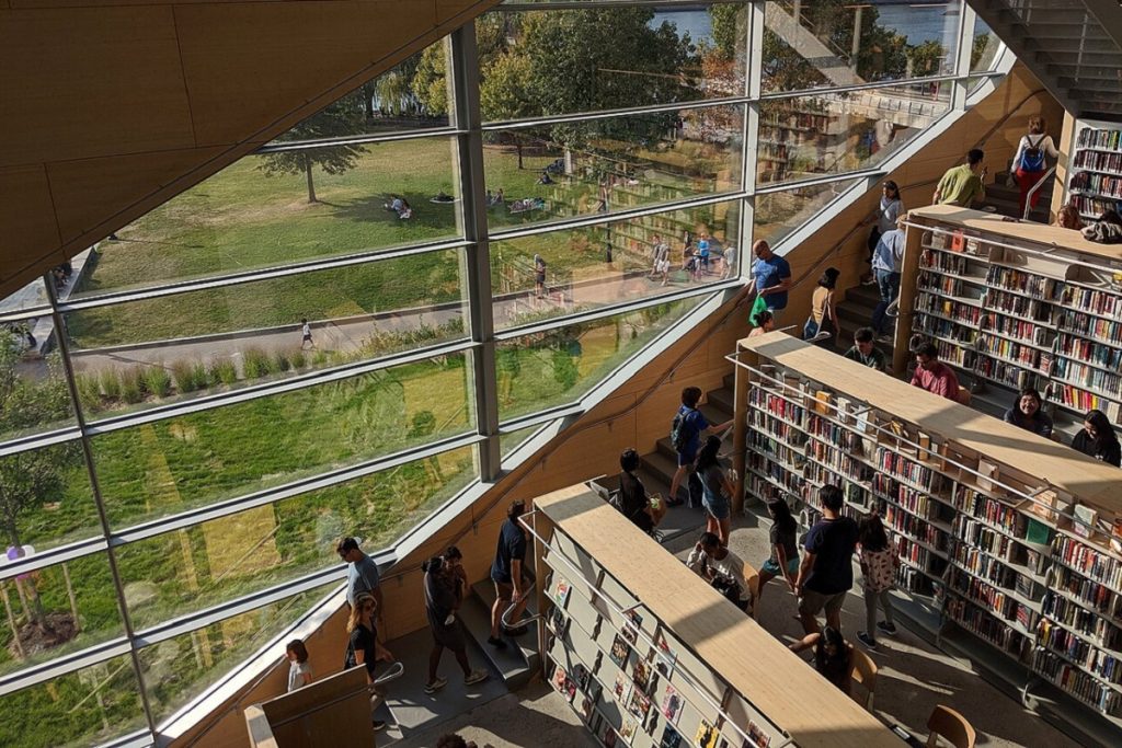Vue en plongée de l'intérieur de la bibliothèque de Hunters Point : un grand escalier en bois longe une façade entièrement vitrée donnant sur un parc verdoyant au bord de l'eau. De nombreux usagers de tous âges circulent entre les rayonnages de livres en bois clair disposés en gradins le long de l'escalier.