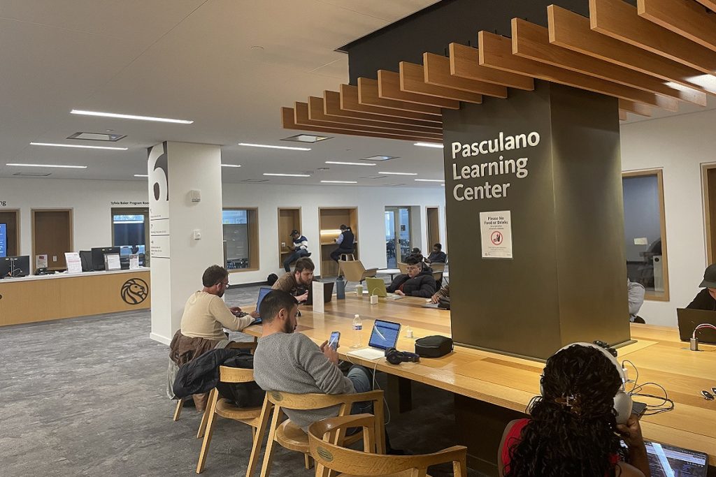 Espace du Pasculano Learning Center : plusieurs usagers sont assis à de longues tables en bois clair, travaillant sur ordinateurs portables ou téléphones. Un pilier sombre porte l'inscription "Pasculano Learning Center". En arrière-plan, un comptoir d'accueil et des salles fermées.