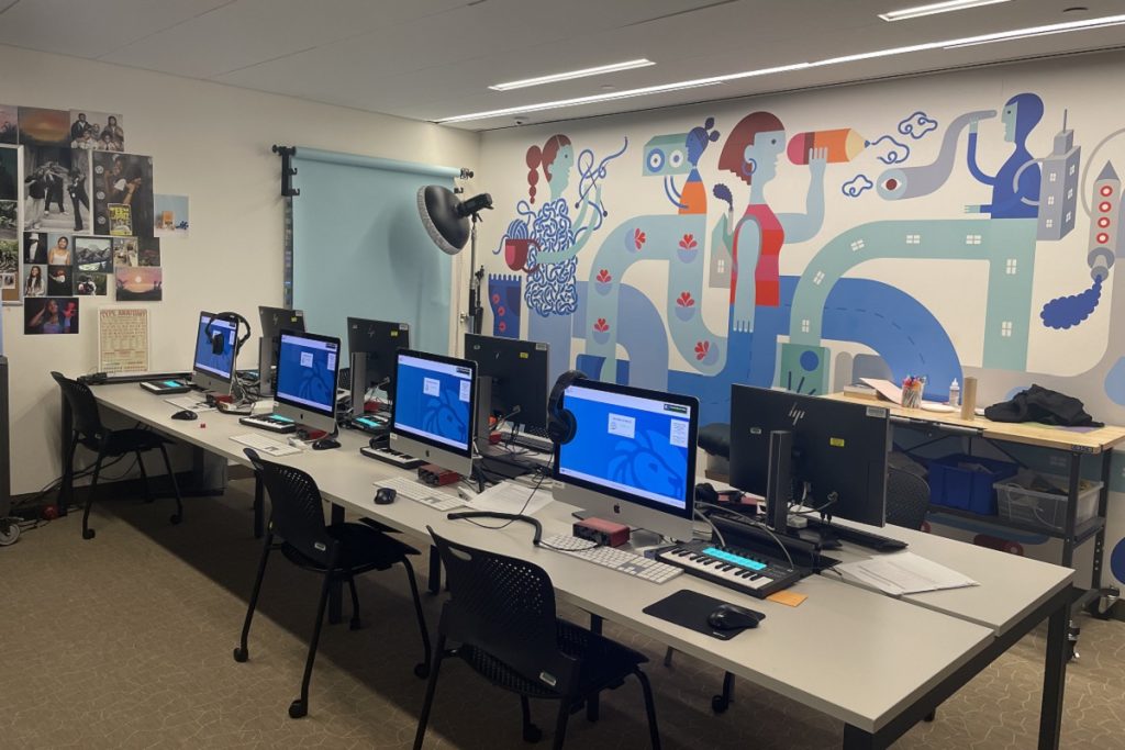 Salle numérique de la Stavros Niarchos Foundation Library : une rangée d'iMac et de claviers MIDI est installée sur une longue table. En arrière-plan, une grande fresque murale colorée représente des personnages stylisés dans un environnement urbain. Un flash photo et des photos de jeunes sont visibles sur le mur de gauche.