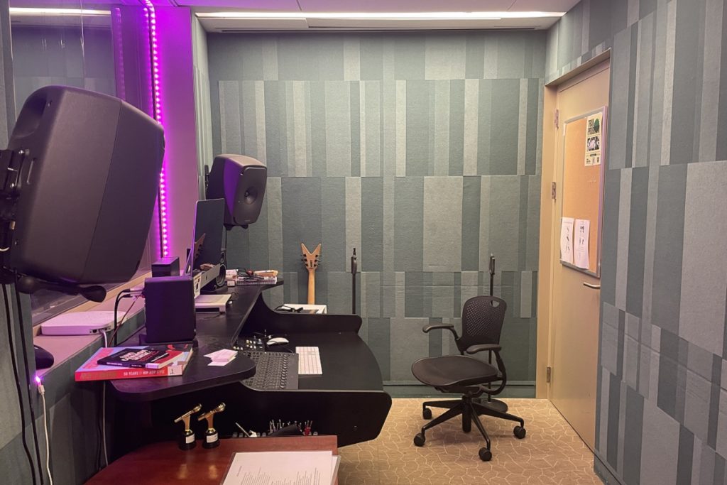 Studio d'enregistrement de la Stavros Niarchos Foundation Library : une petite pièce insonorisée avec des panneaux acoustiques gris sur les murs. Une table de mixage noire, deux enceintes de monitoring et une guitare sont installées à gauche, baignées d'une lumière violette. Une chaise fait face au mur traité acoustiquement. Un panneau d'affichage liège est visible près de la porte.