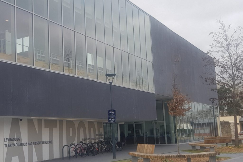 Façade extérieure du bâtiment Antipode, architecture en verre et béton, avec parking à vélos et mobilier urbain en bois.